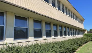 Tan-colored brick building with many windows; bushes in front.