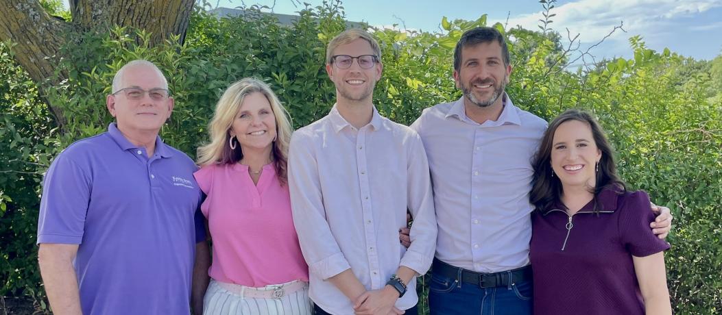 Five people standing next to each other with greenery in background