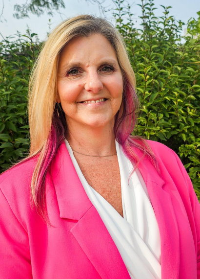 Headshot of woman wearing a white blouse and pink blazer with greenery in background