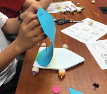A student's hands are shown as he builds a small sailcar (base is corrugated plastic, wheels are wooden beads, skewers are axels, sail is turqoise construction paper)