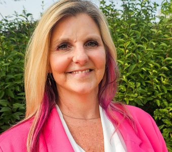 Headshot of woman wearing a white blouse and pink blazer with greenery in background