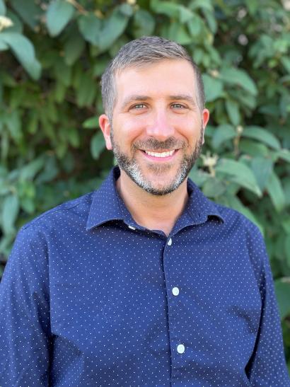 Close-up of man with a beard wearing a blue dress shirt. Leaves and greenery in background.