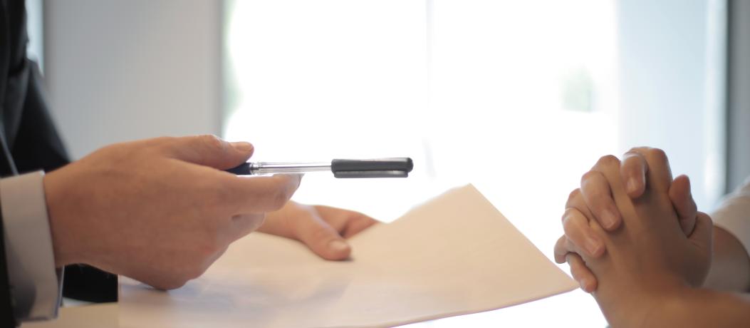 Two people's hands are shown on a desk. One set of hands is holding a paper and pen.