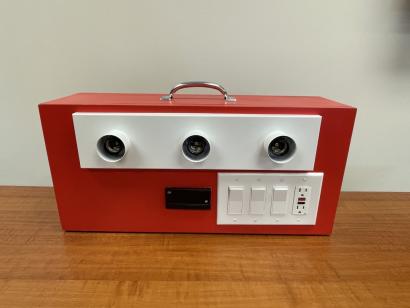 a box on a table that has 3 lightbulbs, 3 switches, a watt meter, and an outlet