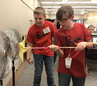 Two boys holding a handheld windmill they made in front of a fun. They are using wind to turn the windmill and lift weights attached on a string.